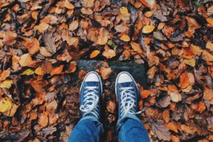 A person has taken a close up picture of their feet while standing on the forrest floor of thick, autumn colored leaves. They are wearing black, old school Chuck Taylor's All-Star, low top sneakers. Their jeans are rolled up just above the ankles. The surrounding leaves are beautiful. The only thing missing in this picture is a pumpkin.