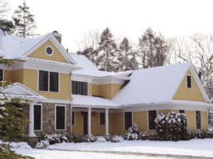 Two story, tan colored house with white trim covered in snow. In the background are snow covered pine trees clearing the height of the house.